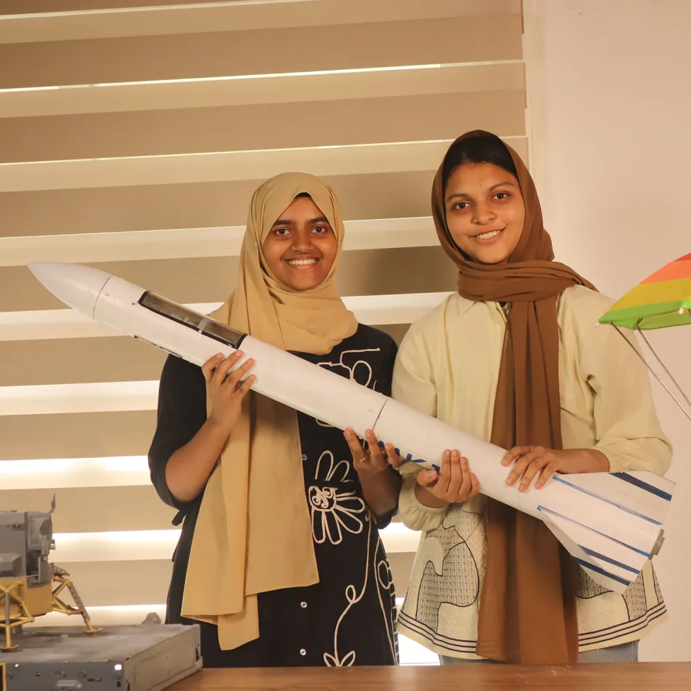 Two students holding a large model rocket they built, beside a parachute recovery model, in a Global AeroSports aerospace lab